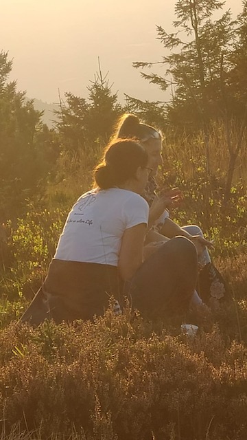 Ein Kräuterwanderführer und ein Teilnehmer sitzen nebeneinander auf einem großen Stein, ihre Silhouetten heben sich vom warmen Glühen der untergehenden Sonne ab. Sie sind in ein intimes Gespräch über Kräuter und das Leben vertieft.