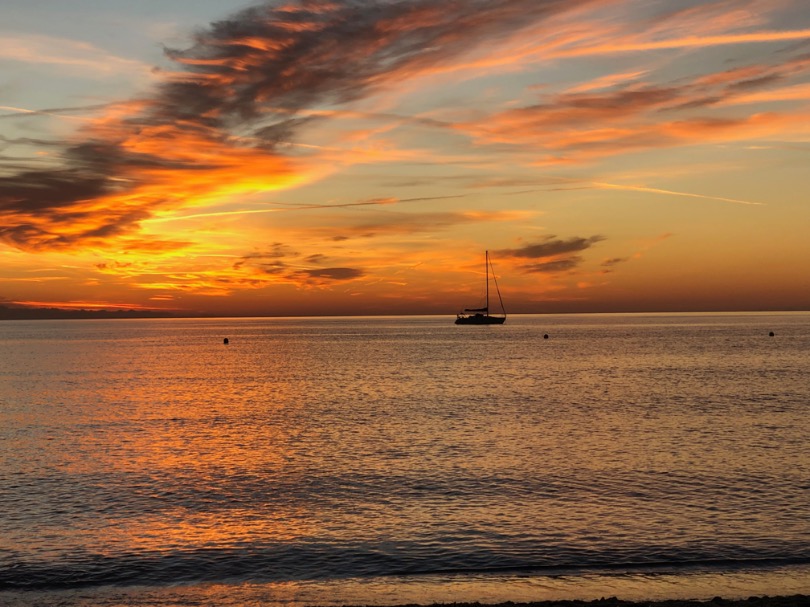 A vibrant sunset over calm waters, with a solitary sailboat in the distance. The sky is painted with hues of orange, pink, and gold, reflecting on the ocean's surface.