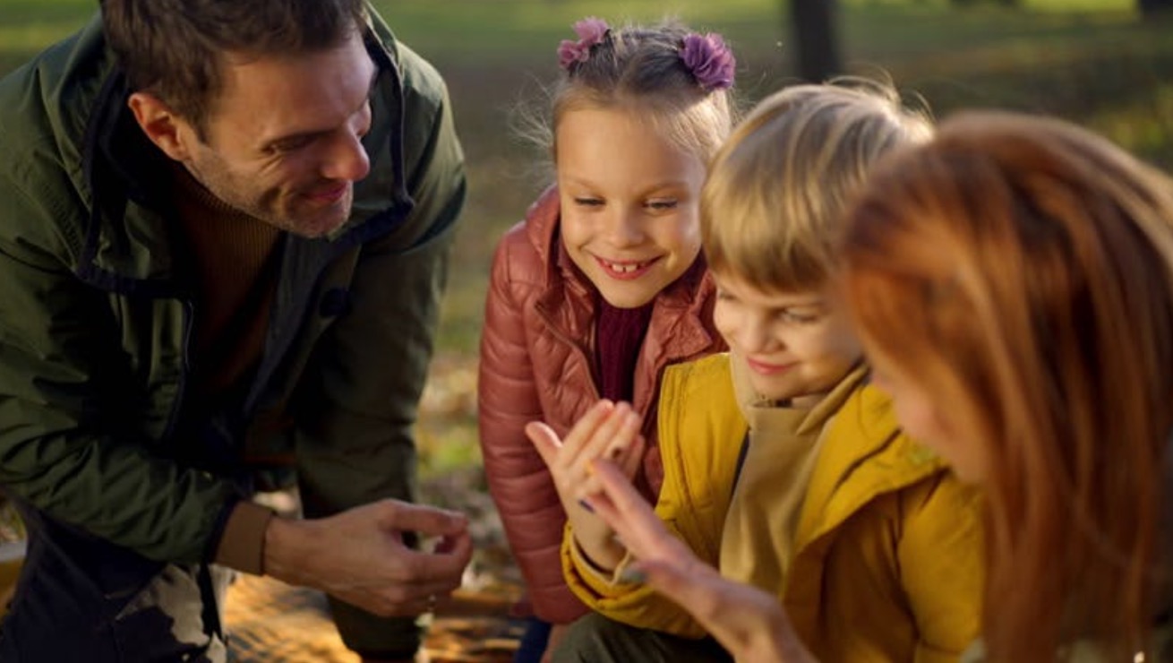 A family outdoors in a park, with two children smiling and examining something in their hands as their parents look on warmly. The scene is full of autumn colors, creating a cozy and engaging atmosphere.