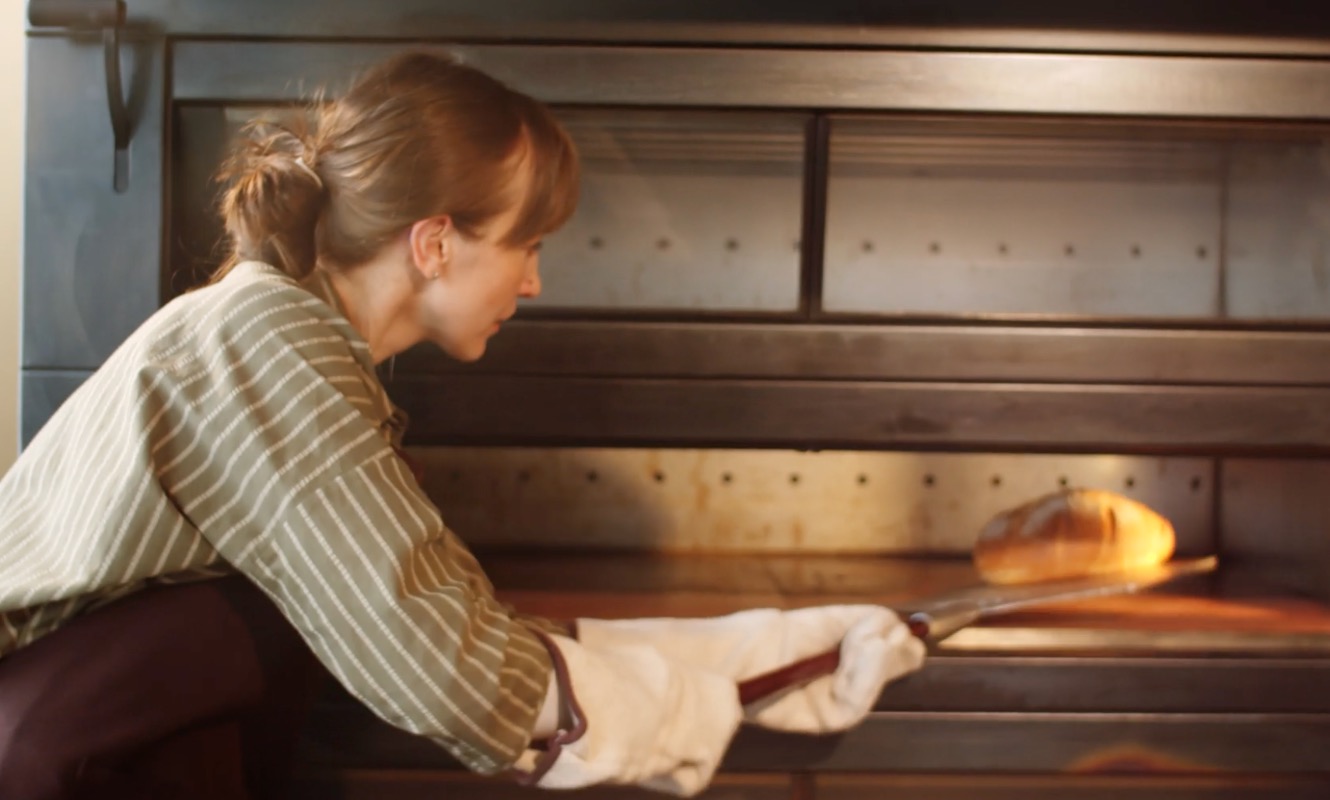 A woman with light brown hair tied back is carefully removing a freshly baked loaf of bread from a traditional oven.