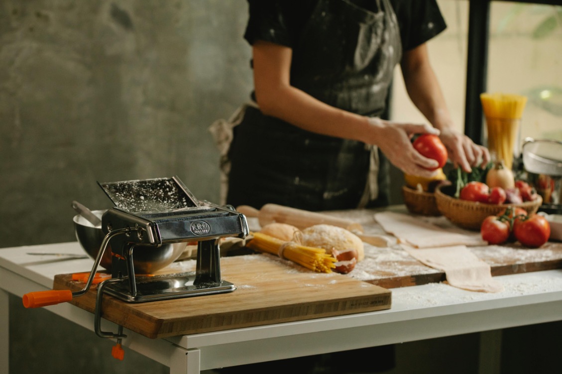 A person in a black apron is preparing fresh pasta in a rustic kitchen. On the table, there is a pasta machine, rolled-out dough, flour, spaghetti, tomatoes, and other fresh ingredients. Warm light is streaming through the window in the background.