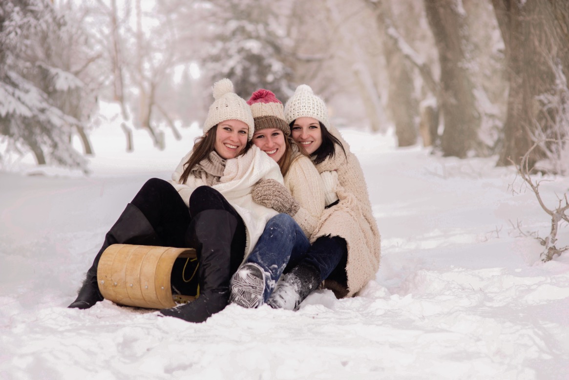 Three young women in winter clothes sit laughing in the snow with a wooden sled. A snowy path stretches behind them.