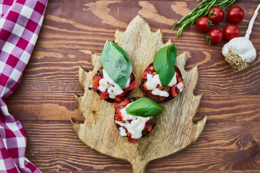 A rustic wooden board with creatively arranged bruschetta, garnished with basil and cherry tomatoes. A red and white checkered napkin lies beside it on a wooden table.