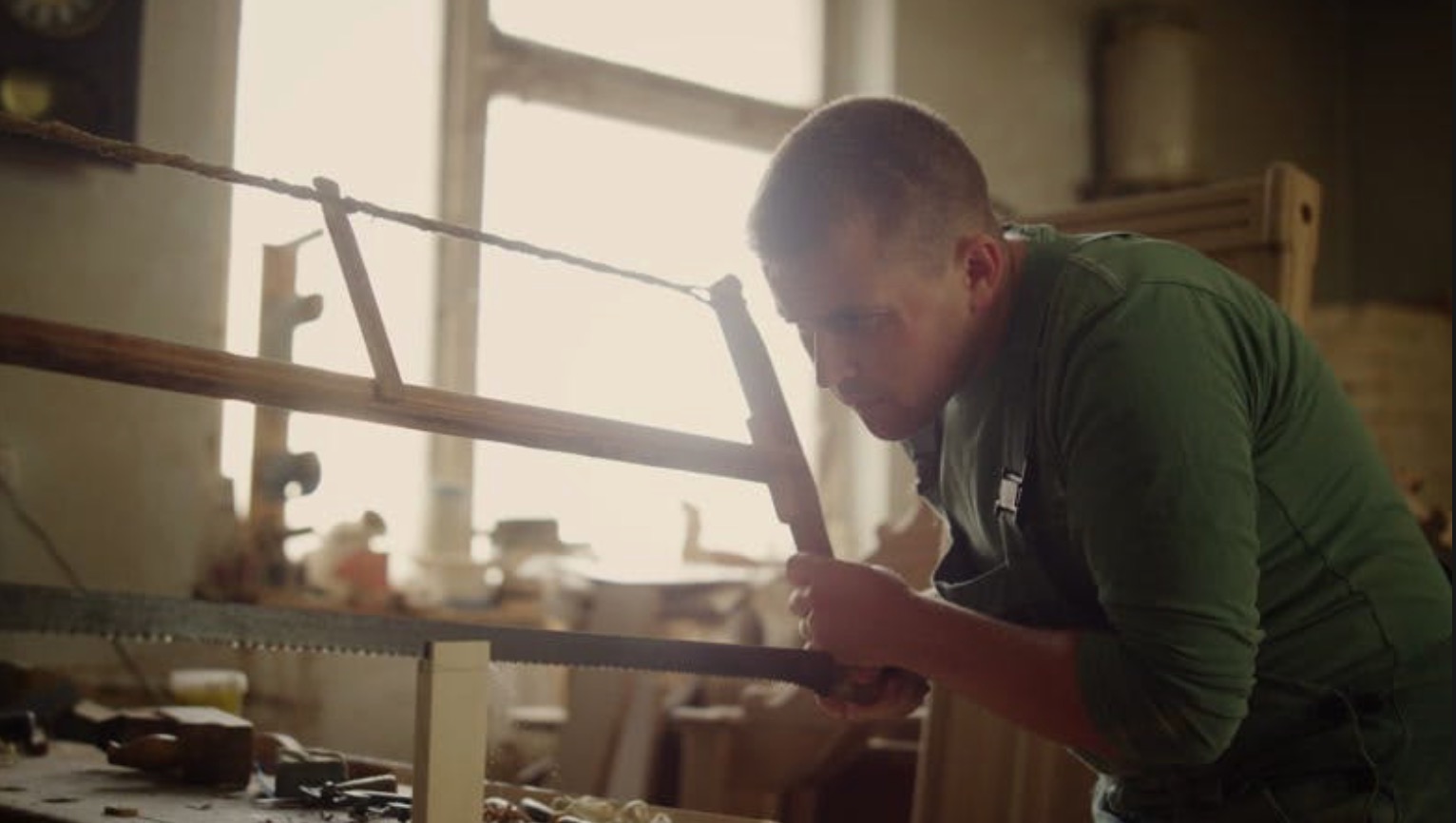 A focused craftsman in a traditional workshop uses a frame saw to cut a piece of wood. The warm, rustic atmosphere of the workshop, with wood shavings and tools in the background, reflects a passion for traditional woodworking.