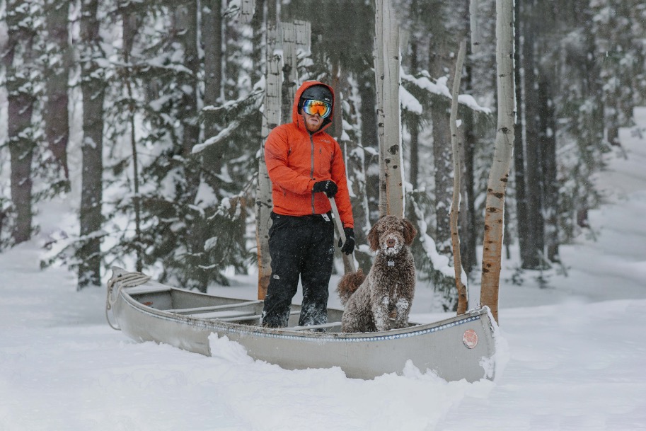 Eine Person in einer roten Winterjacke und Schneeschuhen geht mit einem Hund durch eine verschneite Waldlandschaft. Sie ziehen ein Kanu durch den tiefen Schnee, während es weiter schneit.